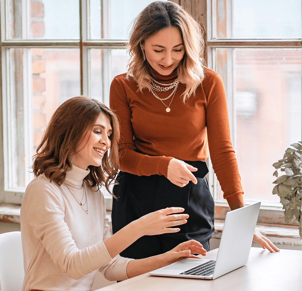 Two women sitting at a table with a laptop computer, and looking at each other